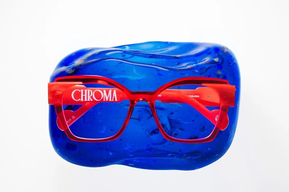 A pair of bright red Etnia Barcelona eyeglass frames with the word “CHROMA” printed on the side, displayed against a vivid blue textured glass background.