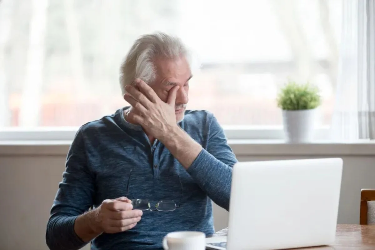Older man rubbing his eyes while using a laptop at a table.