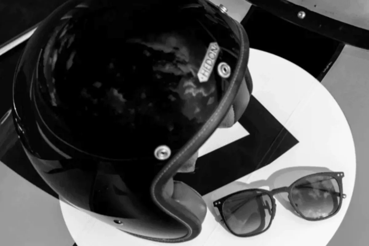 Black motorcycle helmet with reflective visor resting on a white round surface beside sunglasses.