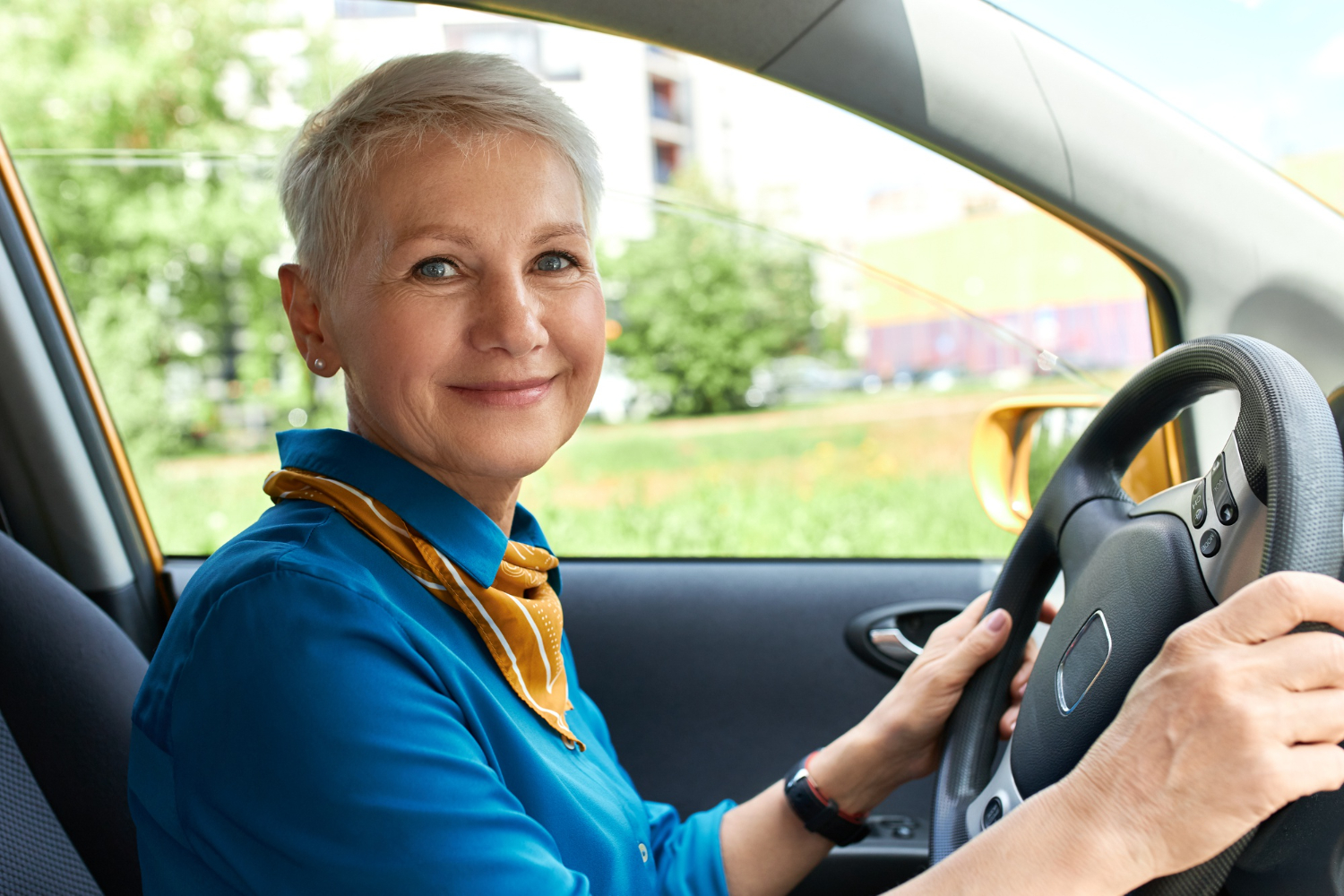 Smiling woman with short gray hair sits in a car, hands on the steering wheel, blue shirt and orange scarf visible in the bright daytime interior.